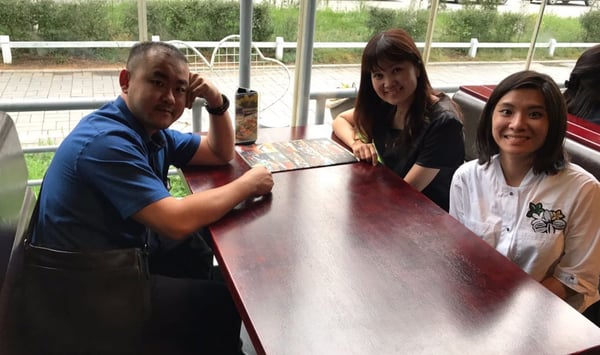 Image shows a man and two women sitting in a booth and looking into the camera